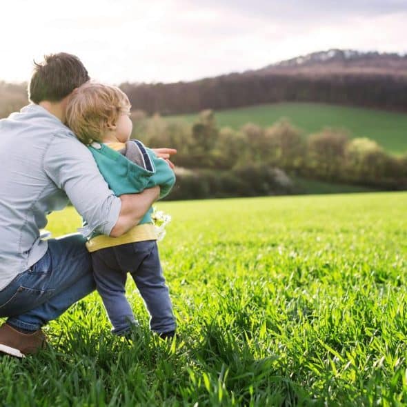A father crouches in a green field holding his toddler.