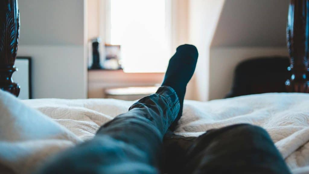 A person’s legs in jeans and socks stretched out on a bed toward a sunlit window.