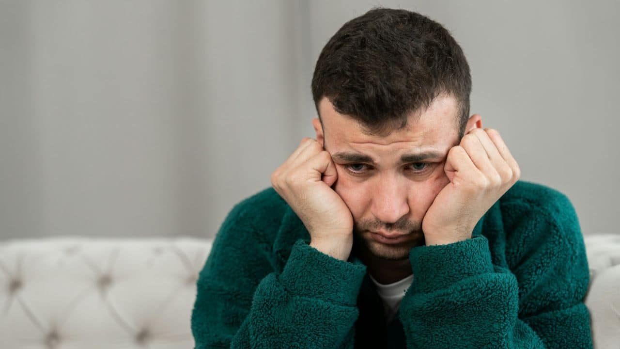 A distressed man in a green robe sits with his face in his hands.