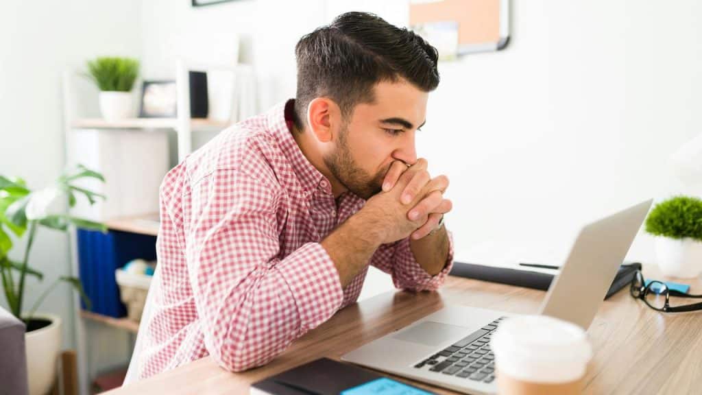 A man in a checkered shirt leaning on his desk while looking at a laptop.
