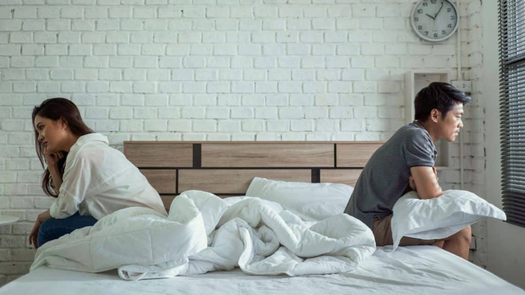 A man and woman sit on opposite sides of a bed, looking away from each other.