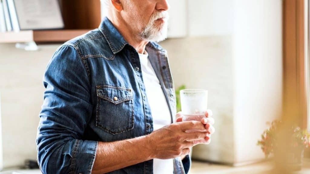 A bearded man in a denim shirt holds a glass of water.