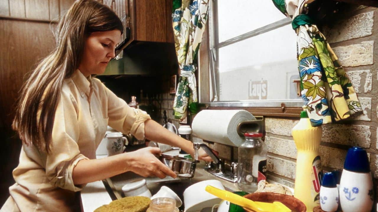 A woman with long brown hair stands at a kitchen sink, washing dishes.