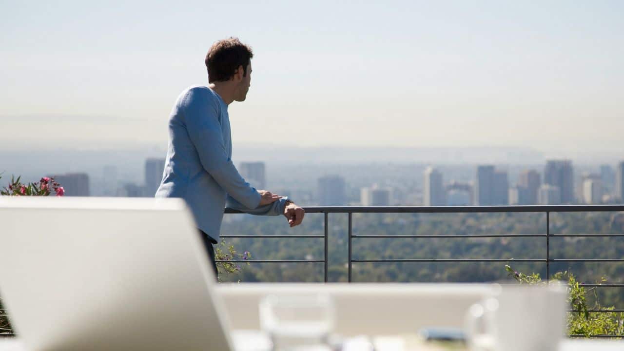 A man in a light blue shirt leans on a balcony railing, looking at a city skyline.