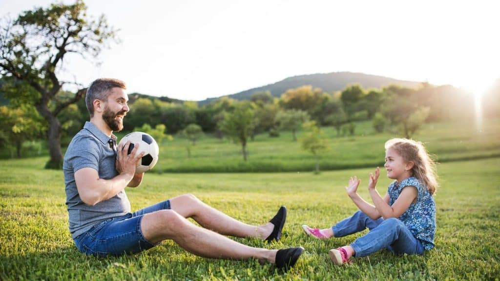 A man holding a soccer ball with his daughter on the grass.