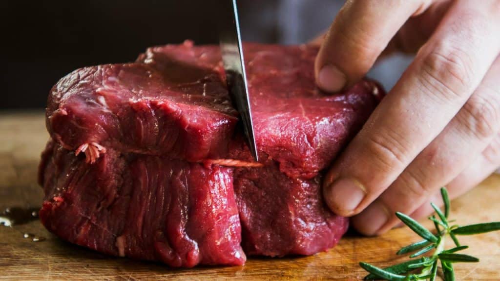 A hand slicing a raw beef roast on a wooden cutting board.