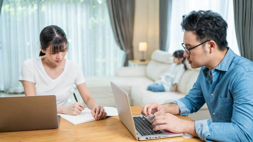 A couple working on laptops at a table while their child sits on a couch behind them.