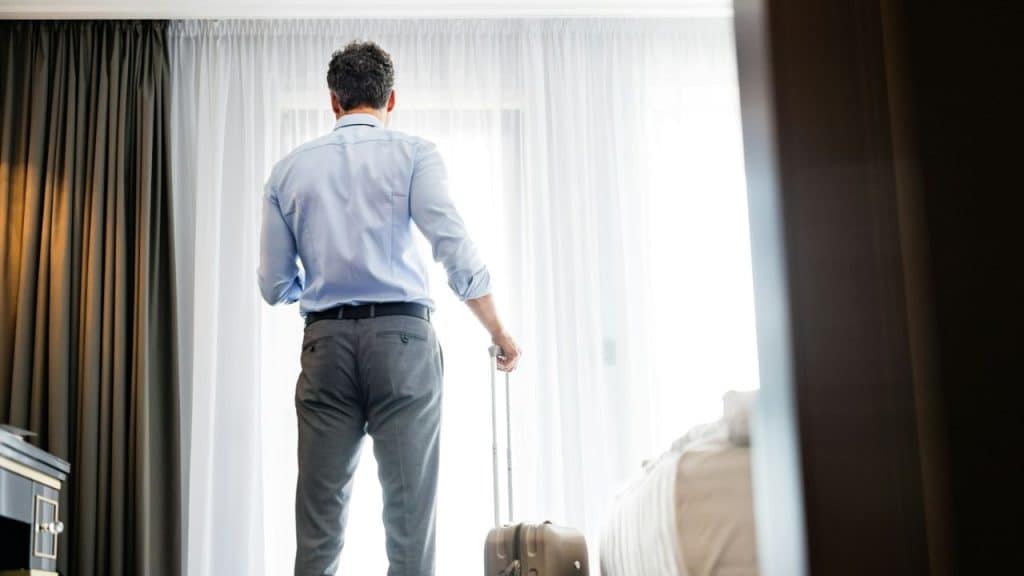 A man with a suitcase stands in a hotel room looking out the window.