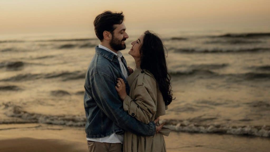A smiling couple embraces on a beach at sunset, looking into each other's eyes.