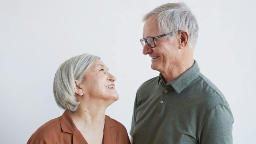 A smiling elderly couple with grey hair looks at each other.