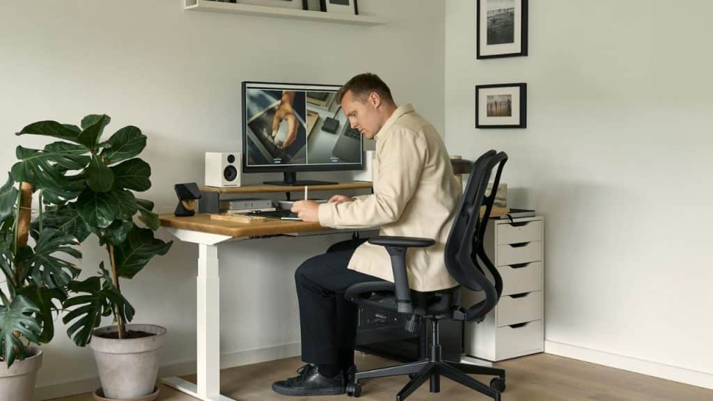 A man works at a modern desk with a computer monitor, plant, and filing cabinet.