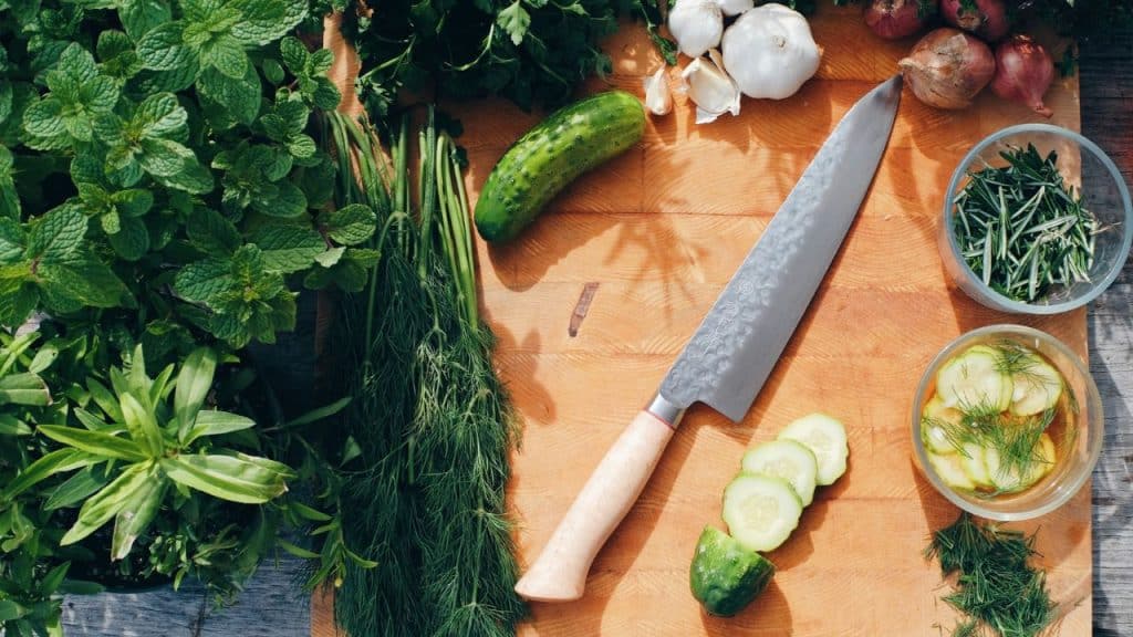 A top-down view of a chef's knife on a wooden board with fresh herbs and vegetables.