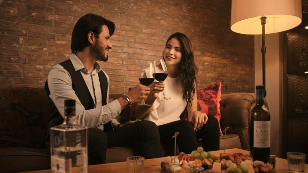A man and woman toast with red wine glasses while sitting on a couch.