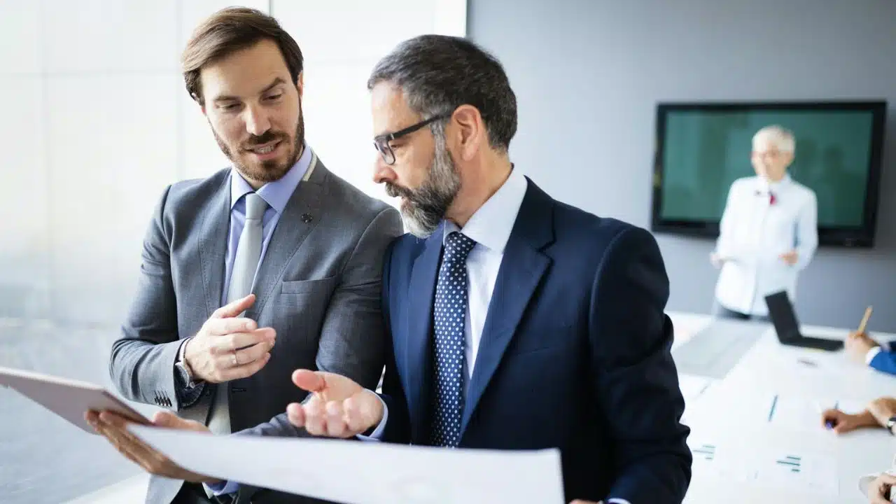 A man in a grey suit looks at papers with a bearded man in a navy suit.