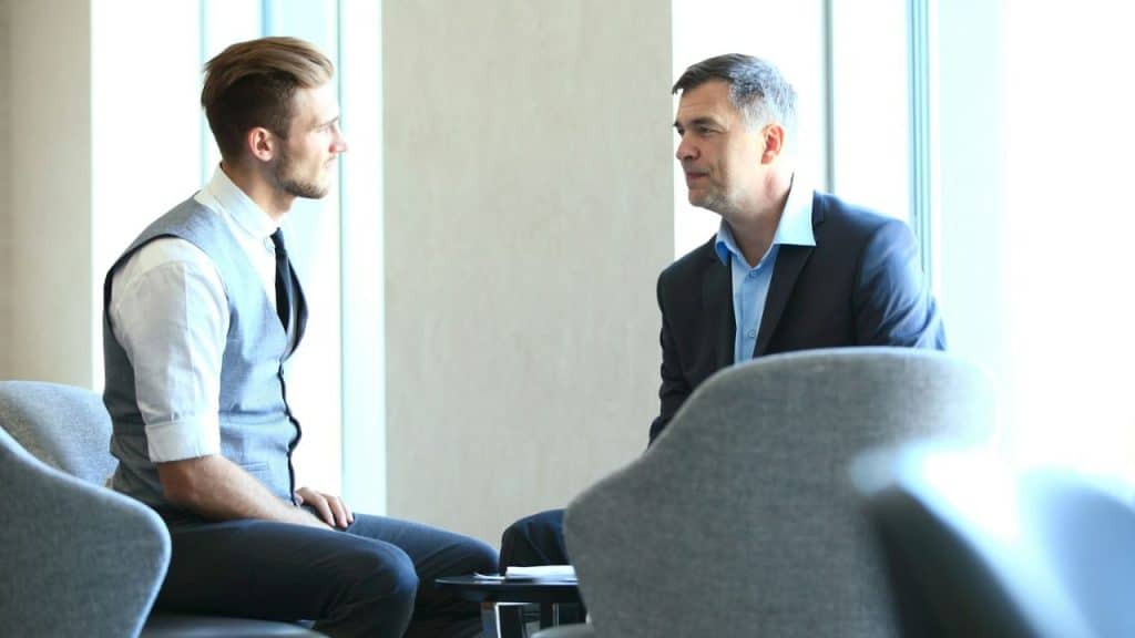 A man in a vest and tie sits facing another man in a suit, both in conversation.