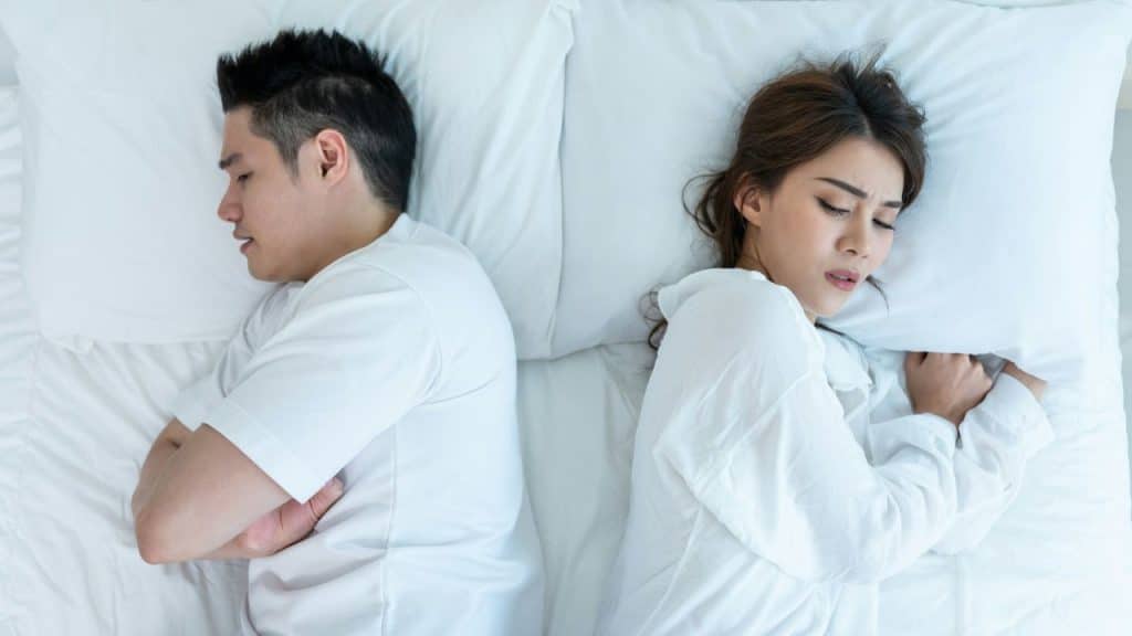 A man and woman lie in bed, back to back, looking distressed.
