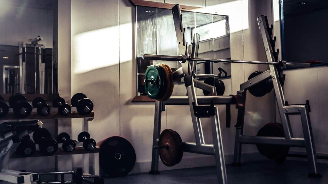 A dimly lit gym interior features weights and a barbell rack, with sunlight streaming in.