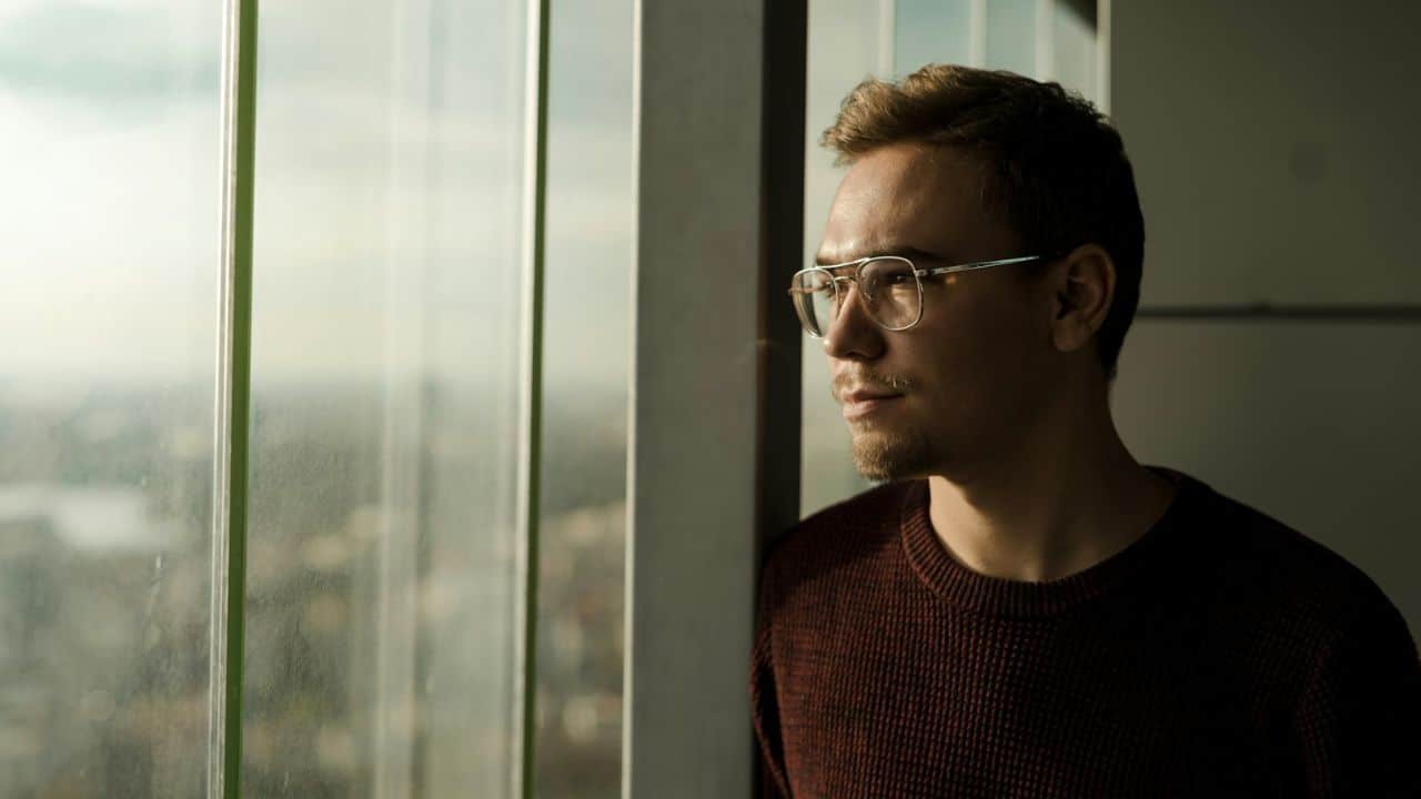 A man in a maroon sweater and glasses looks out a large window.