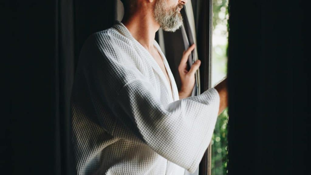 A bearded man in a bathrobe opening curtains.