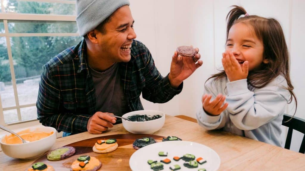 A father offering a decorated cookie to his giggling daughter.