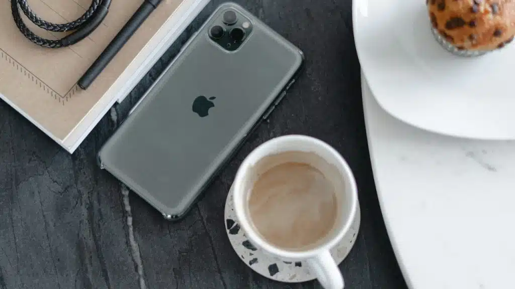 A smartphone, notebook, pen, coffee, and muffin on a dark table.