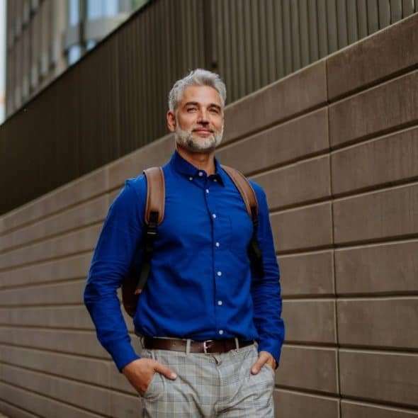 A stylish, gray-haired man in a blue shirt and backpack smiles outdoors.