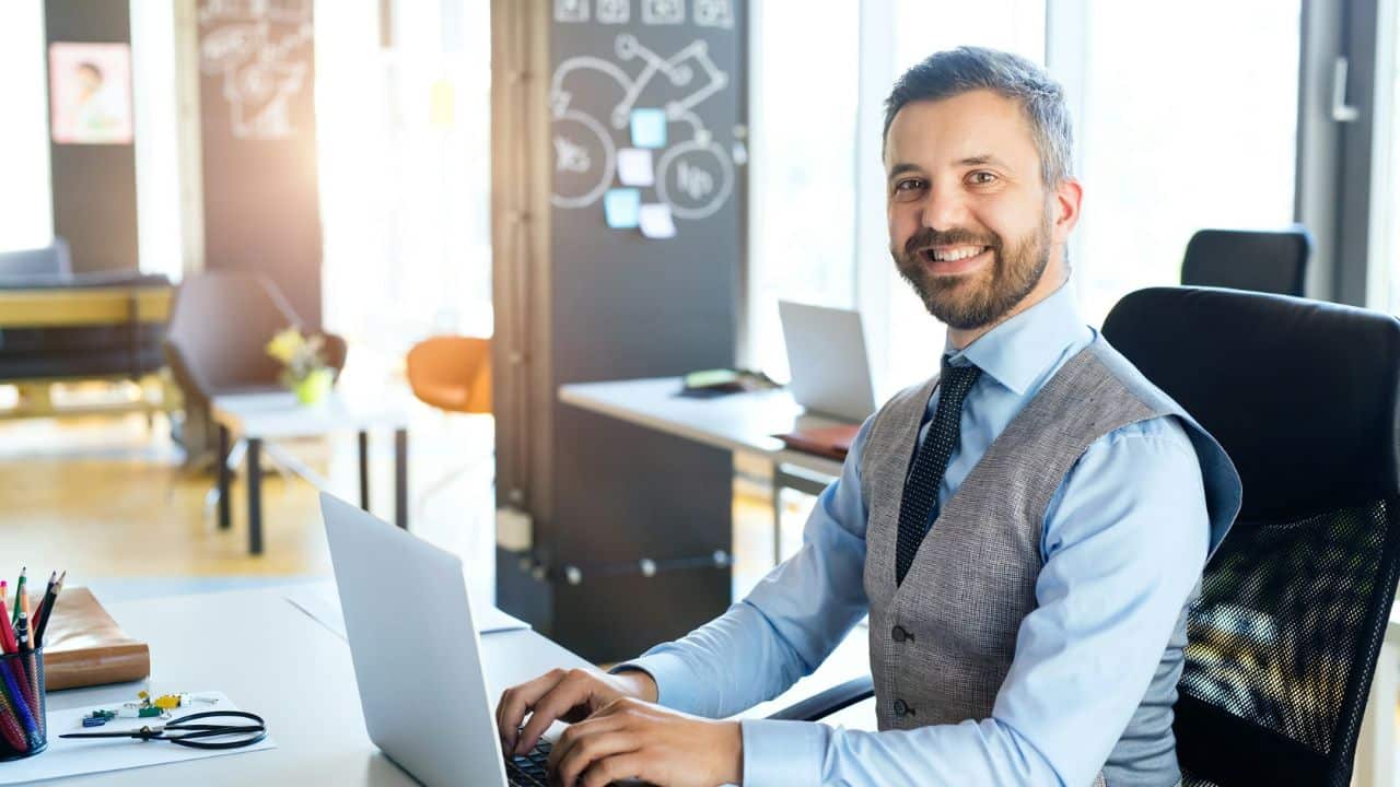 A smiling man in a vest and tie works on a laptop in an office.