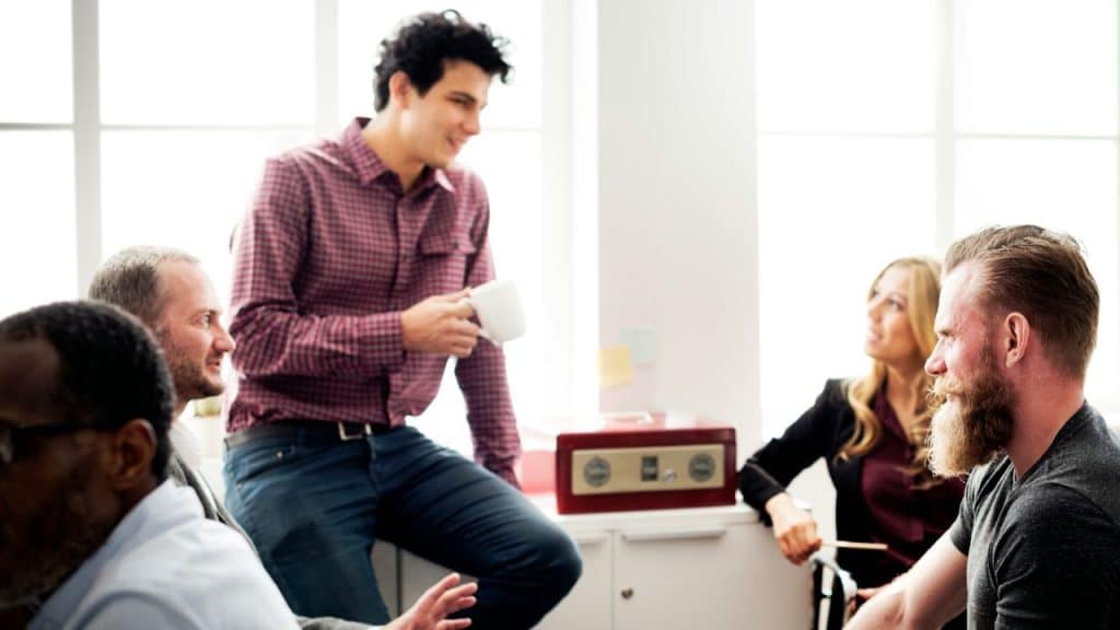 A man holding a mug casually converses with a diverse group of people in an office.