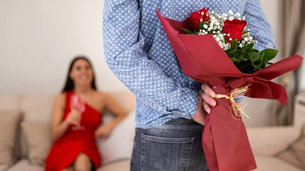 A man hiding a bouquet of red roses behind his back for a smiling woman.