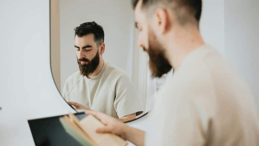 A bearded man reading a book in front of a mirror.