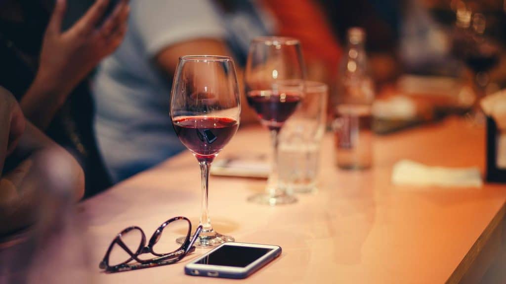 A wine glass and eyeglasses beside a smartphone on a bar counter.