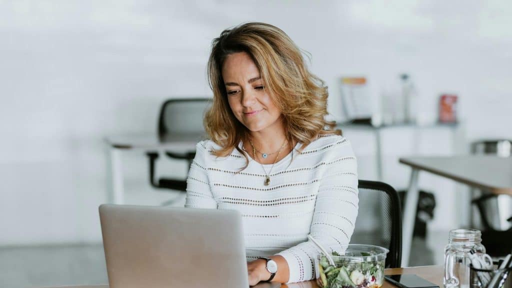 A woman typing on a laptop beside a salad bowl.