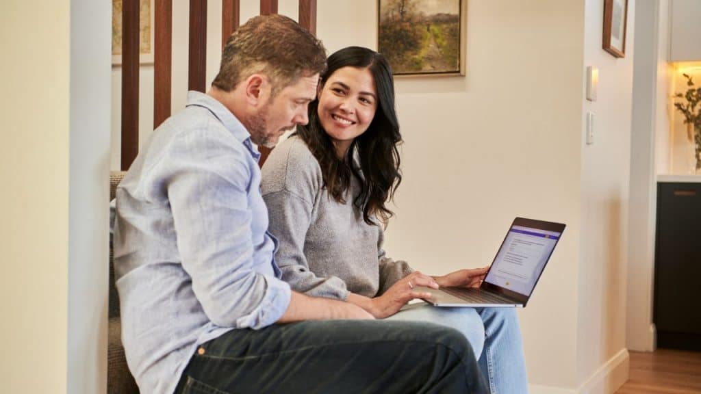 A smiling woman looks at a man while they both look at a laptop screen.