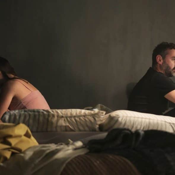 A man and woman sit separately on a bed, facing away from each other.