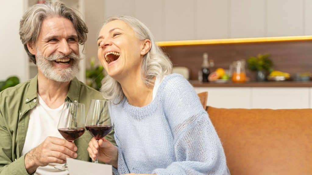 A happy older couple shares a laugh while holding wine glasses on a couch.