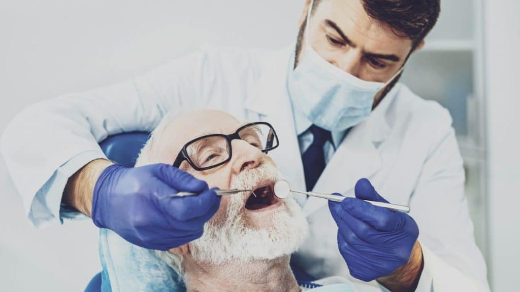 A masked dentist with blue gloves examines an elderly patient's mouth.