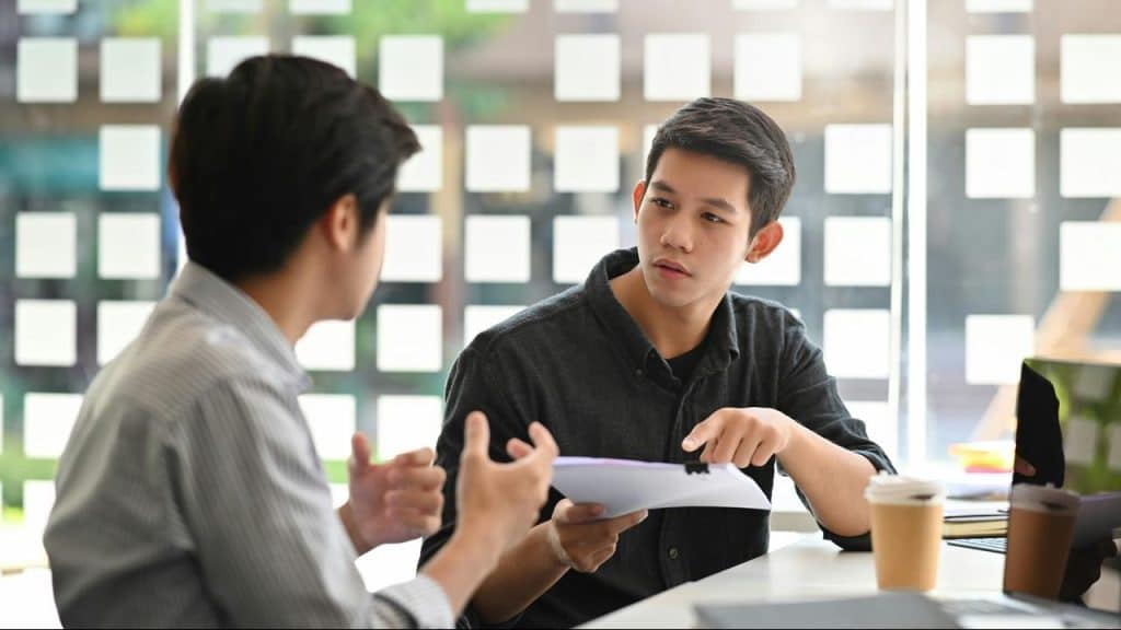 A young man pointing at papers while talking with a coworker.