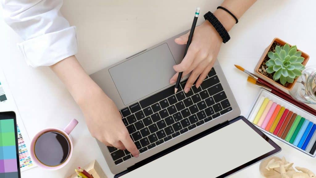 A person using a laptop amid art supplies and a coffee mug.