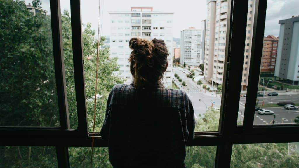 A woman with a bun looks out a window at a city with tall buildings and trees.