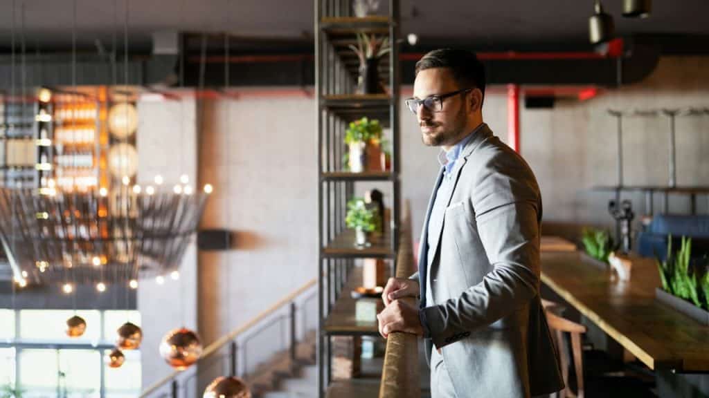 A man in a suit and glasses stands by a railing, looking out over a modern interior.