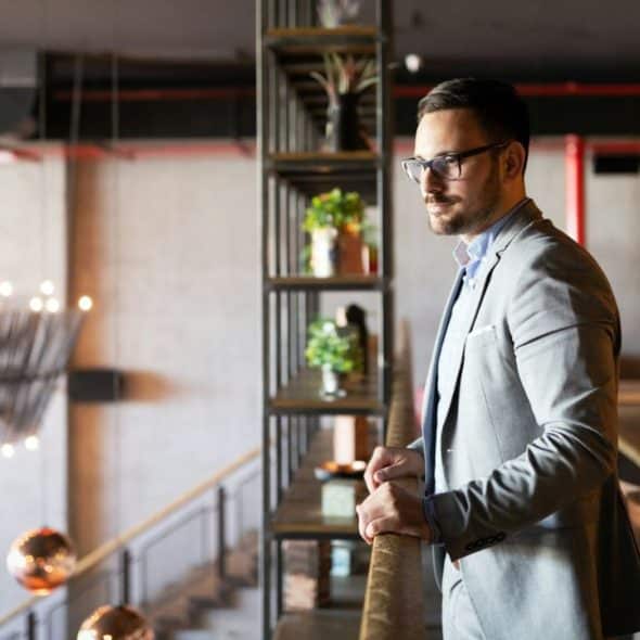 A man in a suit and glasses stands by a railing, looking out over a modern interior.