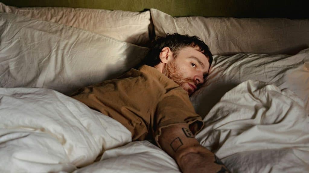 A man with short, dark hair and a beard lies in bed, looking away.