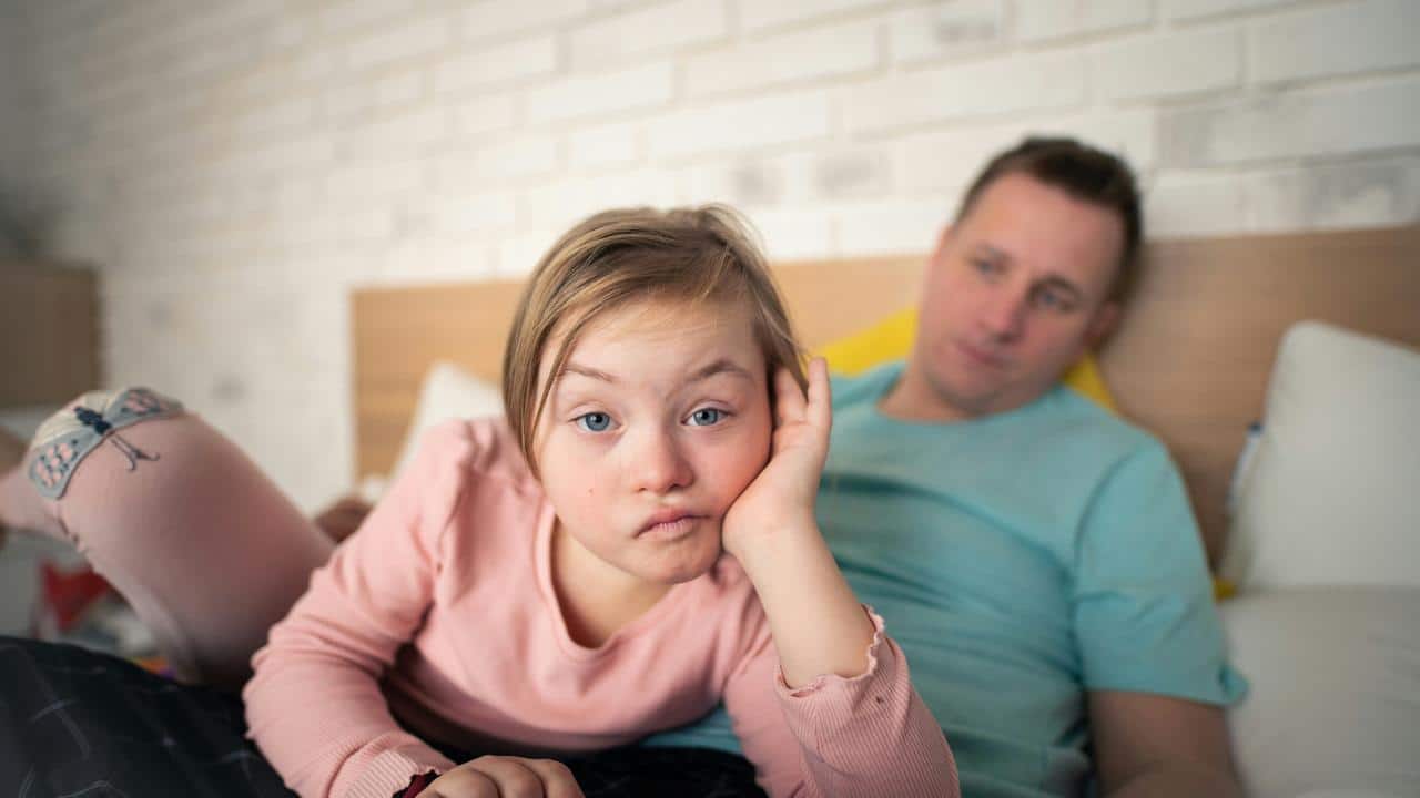 A girl lying on a bed looking bored with an adult behind her.