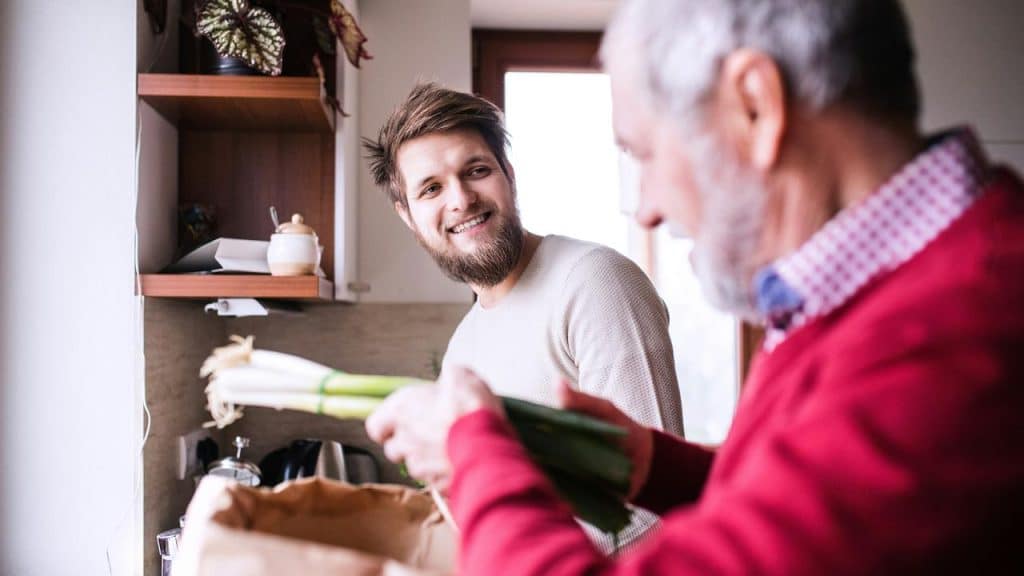 An older man unpacking groceries while a younger man watches.