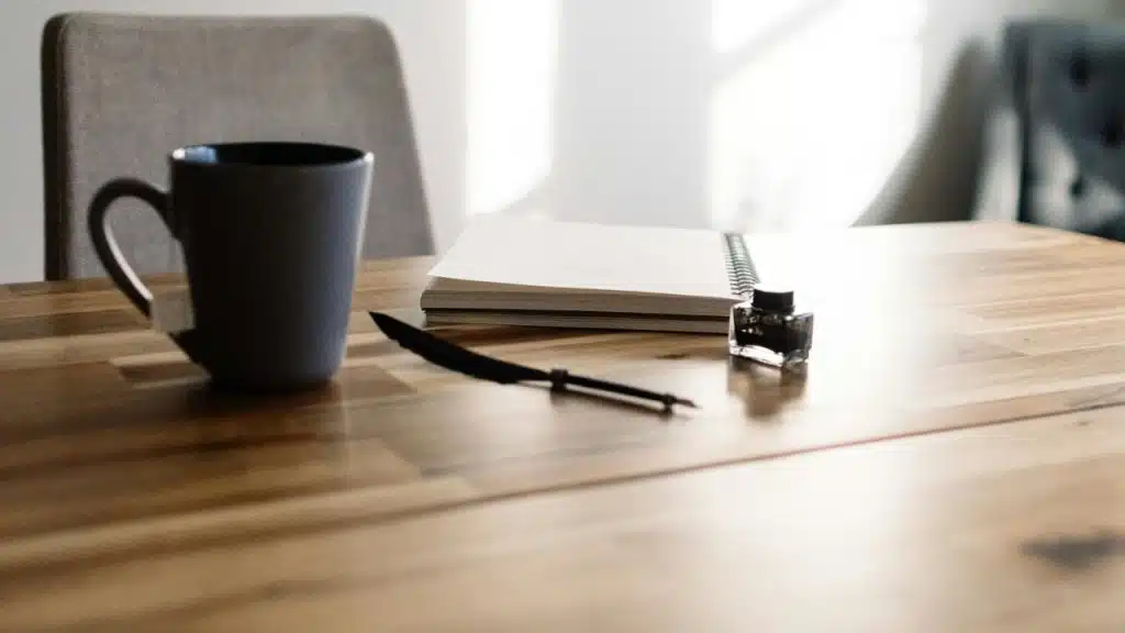 A table with a coffee mug, blank notebook, ink bottle, and quill pen.