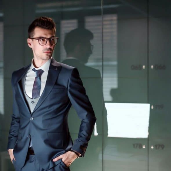 A stylish man in a suit and glasses stands confidently, looking to the side.