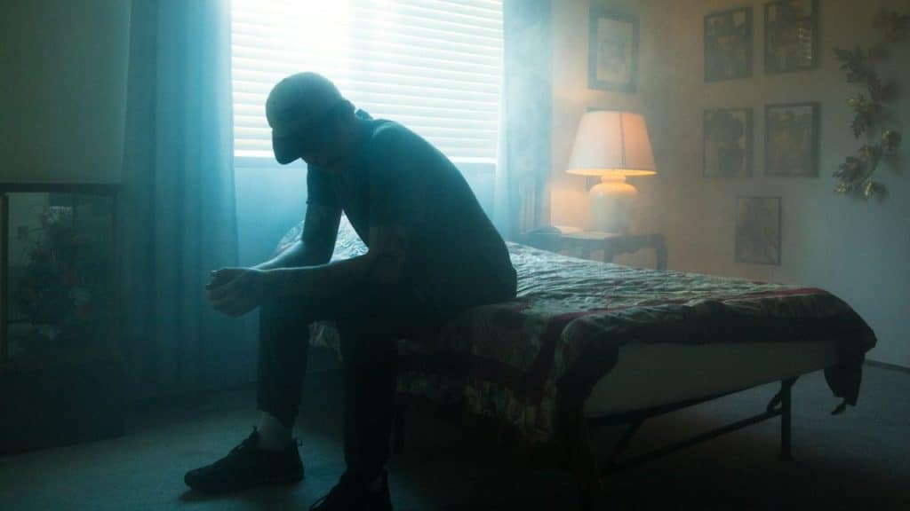 A somber figure sits on a bed in a dimly lit, hazy room, head bowed.