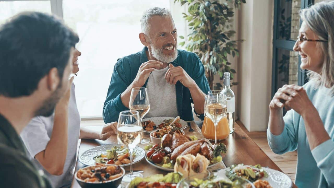 A man with a white beard smiles at a woman across a dining table laden with food and drinks, surrounded by other people.