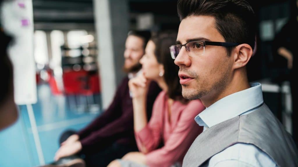 A man in glasses attentively listening during a meeting with colleagues blurred behind him.