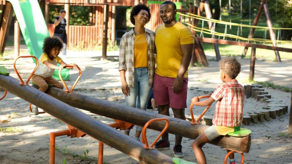 A family watching two children play on a seesaw at a sunny playground.